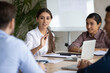 © fizkes - Confident young arab ethnicity woman sitting at table with diverse colleagues, explaining marketing strategy or developing project ideas at brainstorming meeting, office communication concept.