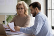© fizkes - Confident focused middle aged 50s businesswoman in eyeglasses discussing marketing research results or company sales statistics paper reports with skilled young male employee at office meeting.