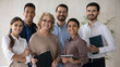 © fizkes - Portrait of smiling 60s middle aged mature female mentor standing in office with motivated skilled young mixed race staff of international company, happy diverse business people looking at camera.