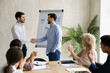 © fizkes - Smiling confident young company leader shaking hands with new worker, celebrating hiring new colleague with multiracial teammates at meeting. Happy diverse coworkers welcoming newbie in office.