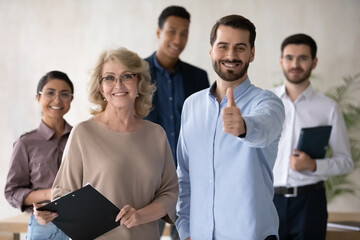 Group portrait of smiling multiracial employees with older senior leader. Motivated young manager showing thumbs up gesture, satisfied with career in international company, looking at camera.