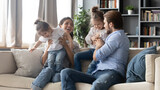 Fototapeta Młodzieżowe - Joyful carefree parents playing with adorable little kids, resting on comfortable couch together at home. Positive happy different generations family having fun with preschool baby daughters indoors.
