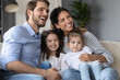 © fizkes - Smiling young married couple relaxing on sofa with adorable little children daughters, enjoying watching TV program, kids show or cartoons together at home, happy family hobby weekend pastime.