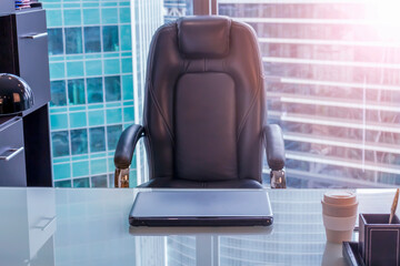 View of empty workplace in office. The chair sits next to the glass table on which the laptop is. Blurred office buildings are visible in the background.