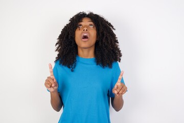 Wall Mural - Young african woman with curly hair wearing casual blue shirt over white background amazed and surprised looking up and pointing with fingers and raised arms.