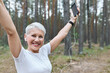 Energetic cheerful retired female with fit slim body posing outdoors in earphones, raising hands, holding cell phone, enjoying music tracks, running. Sports, achievement, goals and determination