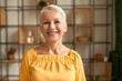 Portrait of joyful stylish middle aged woman with short haircut posing indoors expressing positive emotions, looking at camera with broad happy smile, wearing yellow summer dress, being in good mood