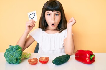 Wall Mural - Young little girl with bang sitting on the table with veggies holding heart reminder scared and amazed with open mouth for surprise, disbelief face