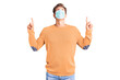 © Krakenimages.com - Young handsome man wearing medical mask amazed and surprised looking up and pointing with fingers and raised arms.
