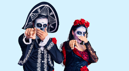 Young couple wearing mexican day of the dead costume over background laughing at you, pointing finger to the camera with hand over mouth, shame expression