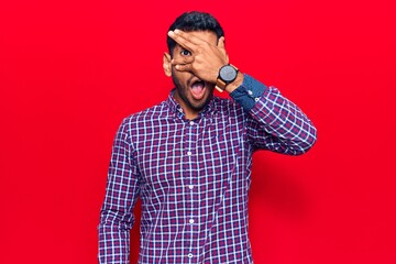 Young latin man wearing casual clothes peeking in shock covering face and eyes with hand, looking through fingers afraid