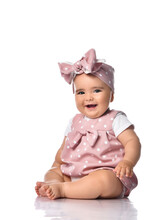 Infant Baby Girl With Opened Mouth Wearing Polka Dot Dress And Headband With Bow Sits On The Floor Looks At Camera