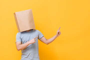 Concept portrait of Anonymous man with head covered with paper bag pointing both fingers up in yellow studio background