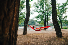 Man Relaxes Lying In The Park On A Hammock Hanging On A Tree On The Background Of The River Embankment And Bridge. A Man Lies On A Hammock In The Park And Has Fun