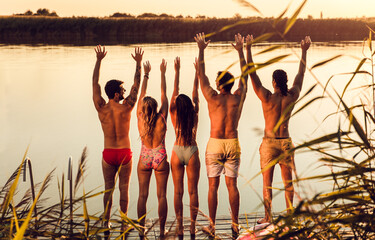 Rear view of group of young friends standing at pier at lake during summer day with arms rise up.