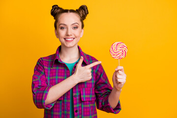 Wall Mural - Portrait of her she nice attractive cheerful girl wearing checked shirt holding in hand demonstrating striped lollypop candy isolated bright vivid shine vibrant yellow color background