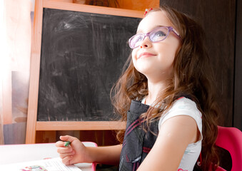 Smart little girl in eyeglasses is sitting at a desk and reading book near empty blackboard. School education concept