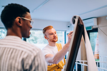 Wall Mural - Young man drawing on flip chart near black colleague