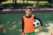 © gorynvd - Happy teen boy in football uniform holds a ball after morning training on the stadium at urban green park.