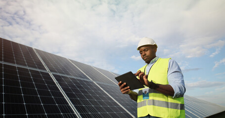 Portrait of african american electrician engineer in safety helmet and uniform using tablet checking solar panels.