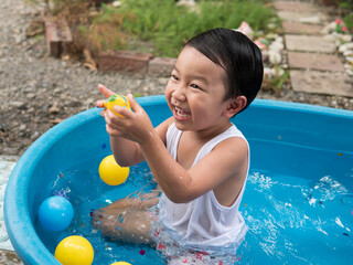 Asian cute child boy laughing while playing water in blue bowl with relaxing face and wet hair in rural nature. Young kid having happy moment in summer. Family activity at home and preschool concept.