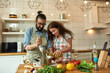 © Svitlana - Italian man, chef cook using hand blender while preparing a meal. Young woman, girlfriend in apron pouring olive oil in the pot, helping him in the kitchen