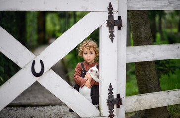 Wall Mural - Small girl with cat standing on farm, looking at camera.