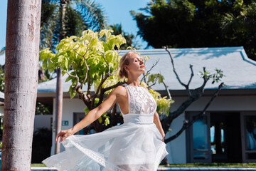 Outdoor portrait of woman in white wedding dress sit near blue swimming pool outside villa in Thailand. Natural bright day light.  