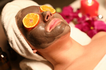 Wall Mural - Woman undergo wellness course and face cleansing. Client lye on massage table with his hair wrapped in towel, coal mask on her face, and orange circles in front of her eyes.