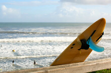 Yellow Surfboard Leaning On A Fence With The Pacific Ocean And Surfers In The Background