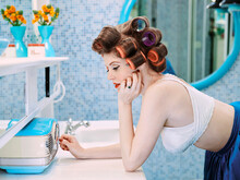Portrait Of A Vintage Styled Woman Turning On Radio In A Mid Century Bathroom, With Hair Curlers In Her Hair.