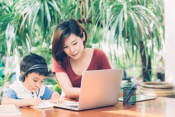 Wall Mural - Young asian mother and son using laptop computer for study and learning together at home, boy writing on notebook for homework and wearing headphone, teacher or mom support child, education concept.