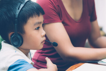 Wall Mural - Young asian mother and son using laptop computer for study and learning together at home, boy wearing headphone for e-learning with distancing, teacher or mom support child, education concept.