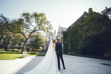 Wall Mural - Elegant bride in a white dress and veil. Handsome groom in a blue suit. Couple near large building