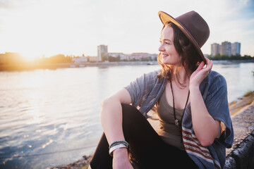 Wall Mural - Portrait of a young beautiful stylish woman in a hat in the city at sunset on the embankment, urban outfit and lifestyle