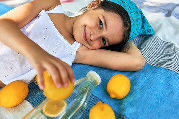 Wall Mural - Gorgeous little girl lying on the blanket with lemons in the park. Pretty kid smiling and having cozy summer picnic with family on sunny day. Child with lemonade resting outdoor.