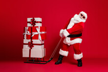 Can't Move. Full Length Photo Of Retired Old Man White Beard Hold Heavy Sled Giftboxes Wear Santa X-mas Costume Coat Sunglass Headwear Leather Boots Isolated Red Color Background