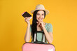 Beautiful woman with suitcase and ticket in passport for summer trip on yellow background. Vacation travel