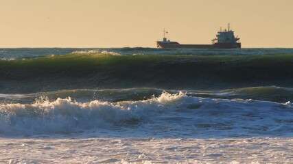 Wall Mural - Sea waves rolling toward beach and sailing cargo ship on sunrise
