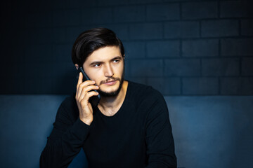 Canvas Print - Portrait of young thoughtful man, talking on the smartphone, sitting on sofa. Background of black brick wall.