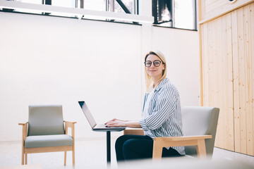 Smart cheerful female employee typing on laptop at workspace