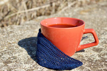 Red cup with coffee and a blue scarf on an old stone on a background of gray dry grass at a halt during a hike. Gourmet rest and coffee on a hike. Gray blurred background, stone texture