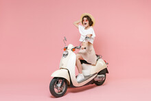 Full Length Portrait Of Excited Young Brunette Woman Wearing White Summer Clothes Hat Glasses Keeping Mouth Open Looking Aside Sitting Driving Moped Isolated On Pastel Pink Colour Background Studio.