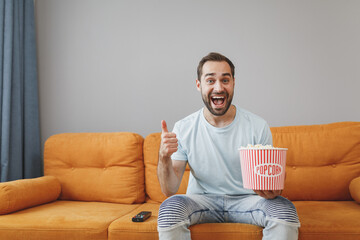 Wall Mural - Cheerful funny amazed young bearded man wearing casual blue t-shirt watching movie film, holding bucket of popcorn showing thumb up sitting on couch resting spending time in living room at home.