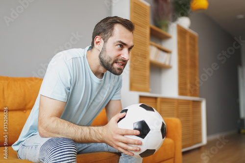 Cuadros en Lienzo Nervous young bearded man football fan 20s wearing basic blue t-shirt cheer up support favorite team with soccer ball looking aside sitting on couch resting spending time in living room at home