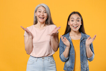 Wall Mural - Surprised excited shocked family two asian women girls gray-haired mother brunette daughter in casual clothes keeping mouth open spreading hands isolated on yellow color background studio portrait.