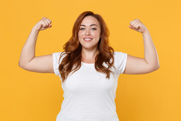 Smiling strong young redhead plus size body positive female woman girl 20s in white casual t-shirt posing showing biceps muscles looking camera isolated on yellow color background studio portrait.