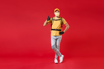 Wall Mural - Full length delivery employee man in yellow cap face mask gloves t-shirt uniform thermal food bag backpack work courier service during quarantine coronavirus covid-19 virus isolated on red background.