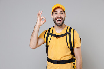 Wall Mural - Delivery employee man guy male 20s in yellow cap t-shirt uniform thermal food bag backpack work courier service during quarantine coronavirus covid-19 virus, posing isolated on gray background studio.