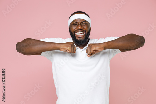 Fotomural Cheerful young bearded african american fitness sports man 20s in white headband t-shirt doing stretching exercising spending time in gym isolated on pastel pink color wall background studio portrait